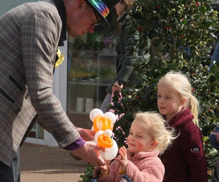 Lange Jan geeft een ballonkunstwerk aan een blij klappend meisje, terwijl andere kinderen op hun beurt wachten
